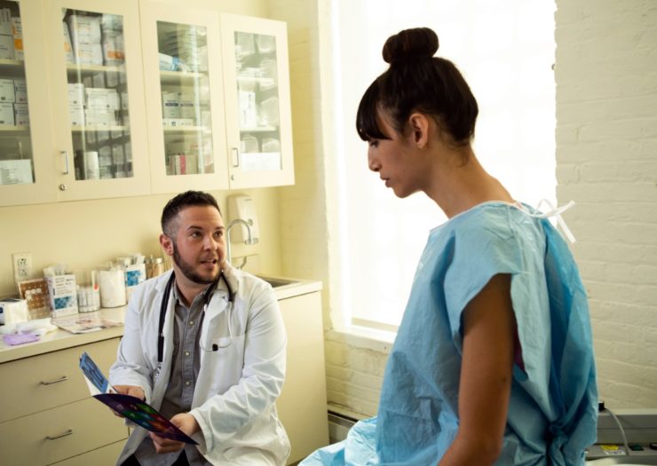 [Photo: A transgender woman in a hospital gown speaking to her doctor, a transgender man, in an exam room.]