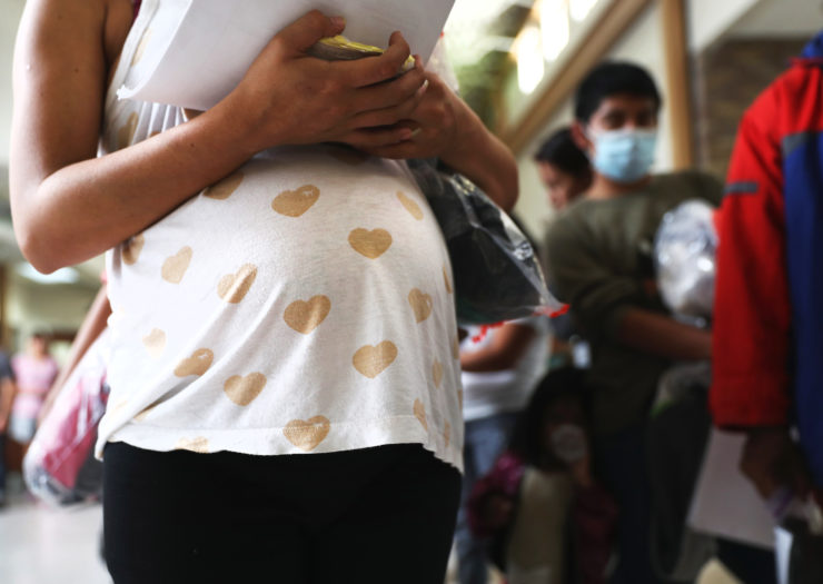 [Photo: A pregnant migrant woman stands in line with other immigrants awaiting inspection.]