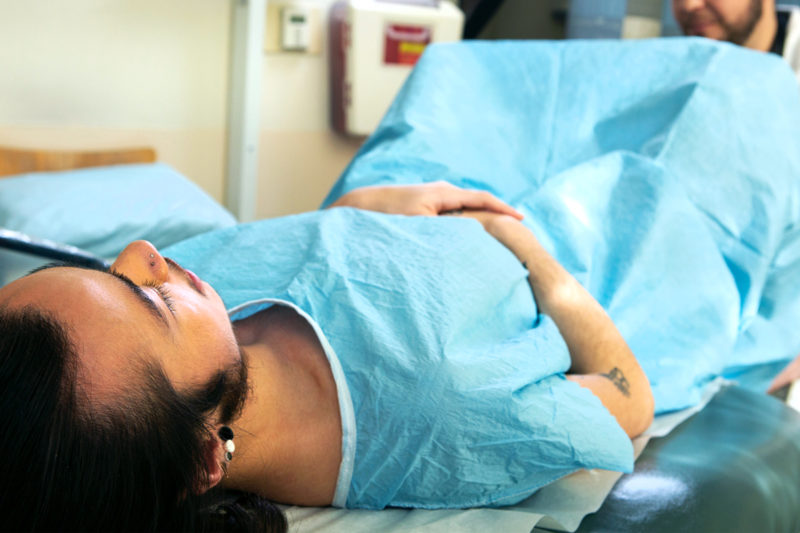 [Photo: A non-binary person wears a gown while they lie down for a pelvic examination.]