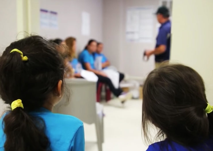 [Photo: Two young, migrant children look on as their mothers wait in a holding room with an officer.]