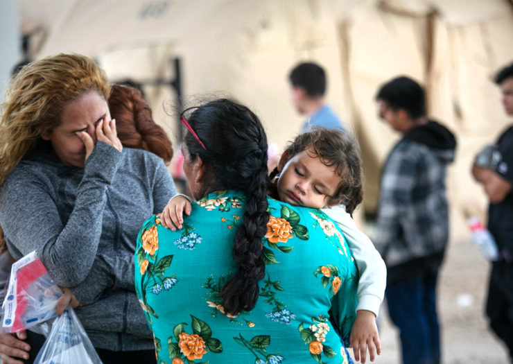 [Photo: In the foreground, two migrant women stand together crying. One woman holds a young chiild. In the background, a group of migrants wait to be interviewed by U.S. Border Patrol agents after they were taken into custody.]