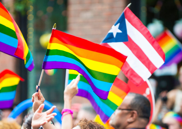 [Photo: A crowd of LGBT rights supporters march as they wave Puerto Rican and rainbow flags.]