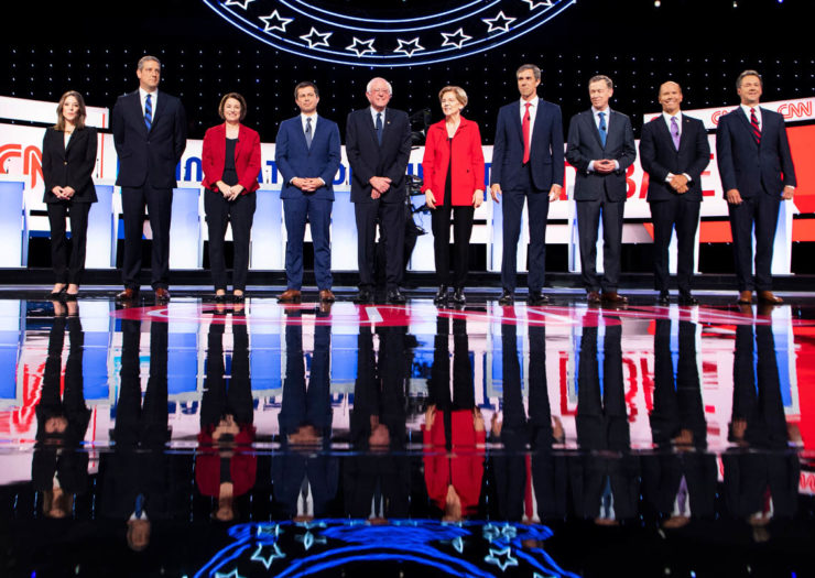 [Photo: Ten of the 2020 democratic presidential candidates stand onstage before the beginning of a debate.]