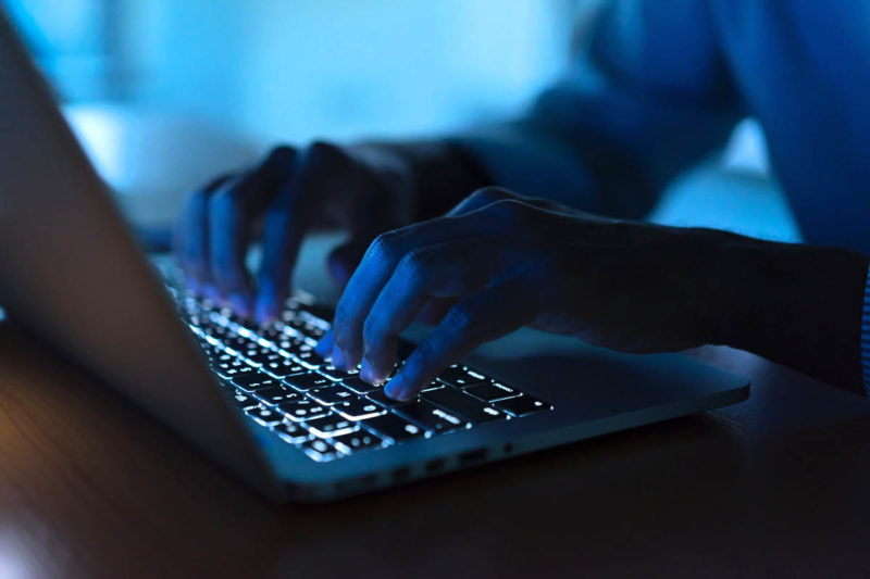 [Photo: A close up of a person's hands as they type on a keyboard in ominous darkness.]