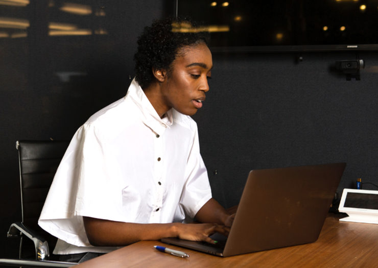 [Photo: A young, Black, gender non-conforming person works on their office laptop late at night.]