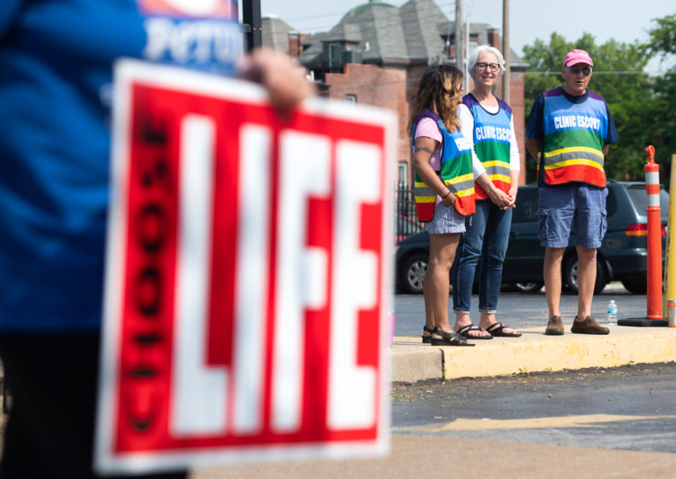 [Photo: Clinic escorts stand in a parking lot as anti-abortion demonstrators hold a protest outside an abortion clinic.]