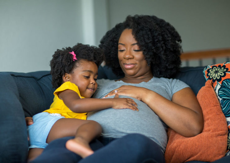 [Photo: A pregnant Black woman reclines on a couch with a toddler touching her stomach.]