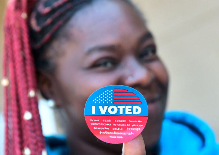 [Photo: A young, Black woman holds up an 'I voted' sticker as she smiles.]