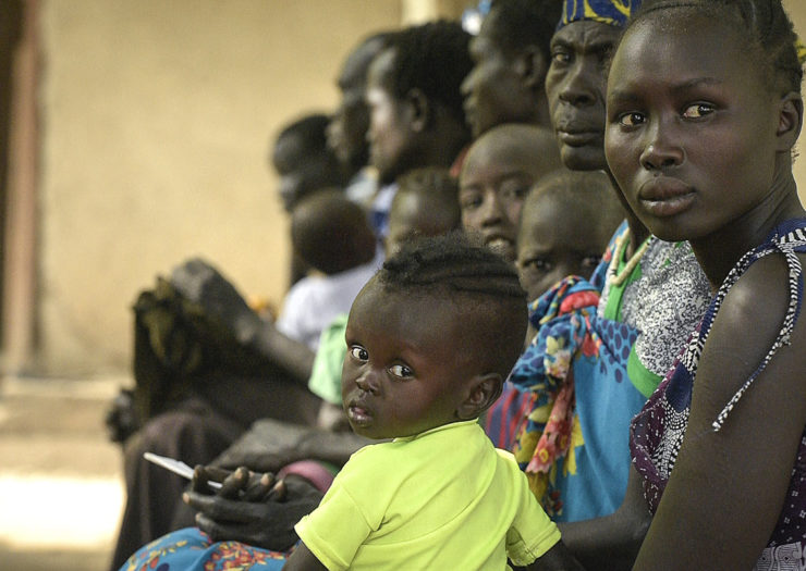 [Photo: An African woman and her baby, and other patients wait for treatment outside a clinic.]