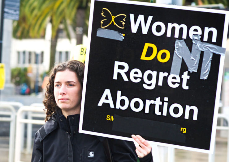 [Photo: A young, white woman holds a sign that reads 'women don't regret abortion'.