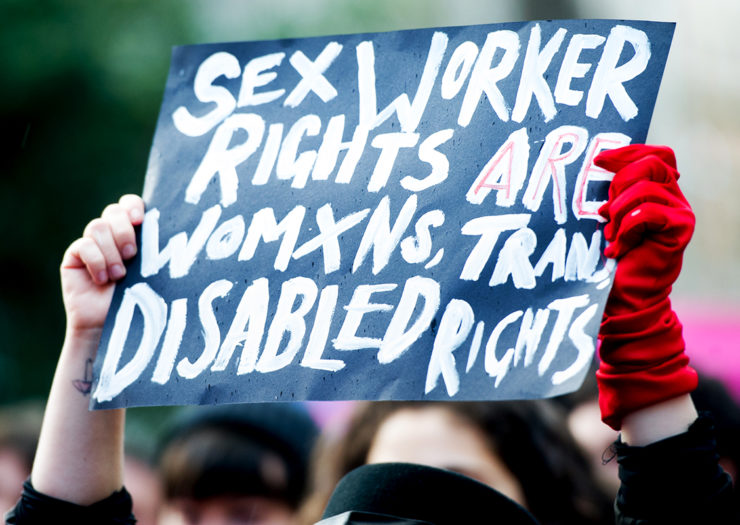 [Photo: A sex worker advocate holds up a sign during a protest. The sign reads 'sex worker rights are womxn, trans, disabled rights.']