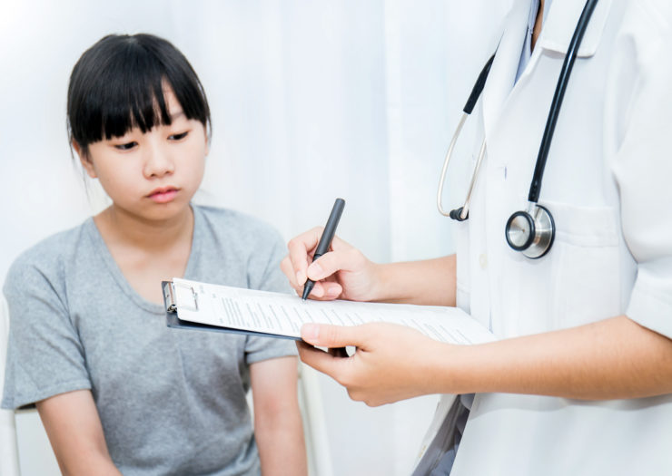[Photo: An Asian teenage patient looks at a clipboard that their doctor is taking notes on.]
