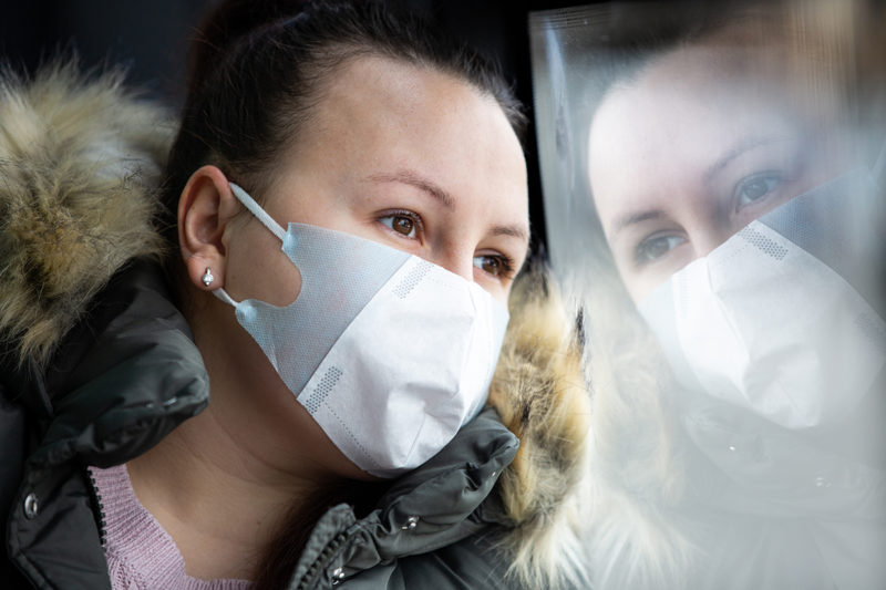 [Photo: A worried woman wearing a face mask looks out the window of a bus.]
