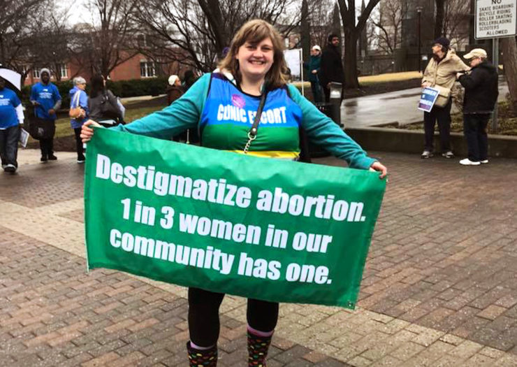 [Photo: An abortion clinic escort smiles as she holds a green banner in support of abortion.]
