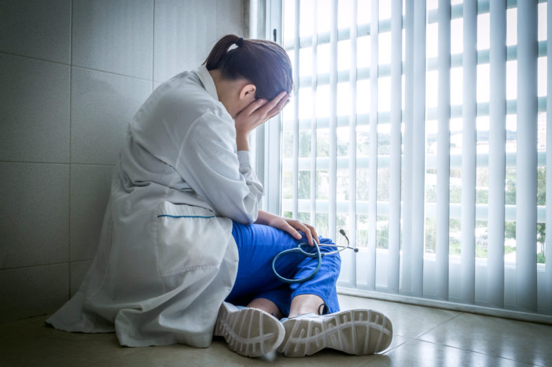 [Photo: A sad and tired doctor holds her head as she sits alone on a hospital floor near a window.]