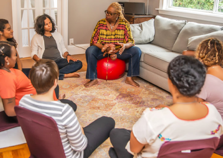 [Photo: A group of predominantly pregnant people of color sit in a living room as they listen to an elder Black birthworker speak.]