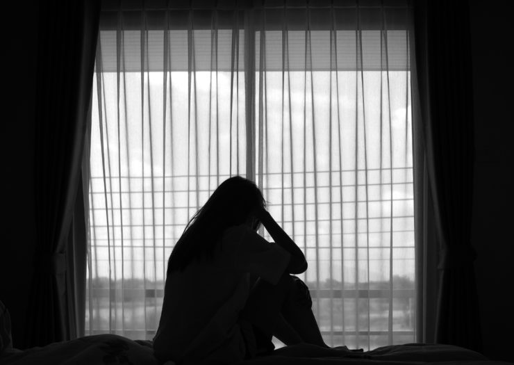 [Photo: A black and white photo of the silhouette of a woman holding her head as she sits on a bed in front of a window.]