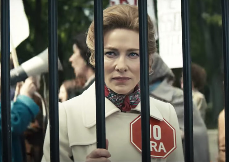 [Photo: An image still of the FX show 'Mrs. America' that shows a white woman scowling and wearing a 'NO ERA' pin in the midst of a protest.]