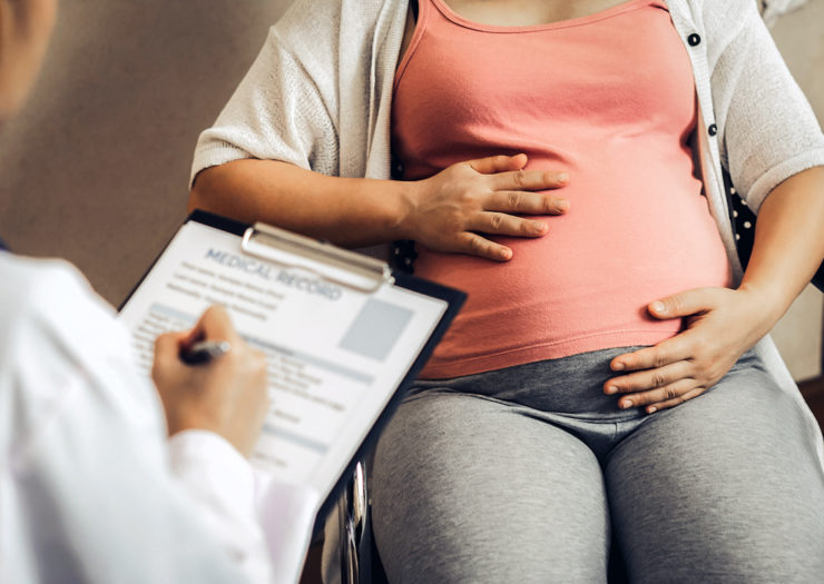 [Photo: A pregnant person holds their belly as they speak to a doctor who holds a clipboard.]
