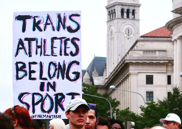 [Photo: A person holds up a sign amidst a crowd in support of trans athletes participating in sports during National Trans Visibility Day.]