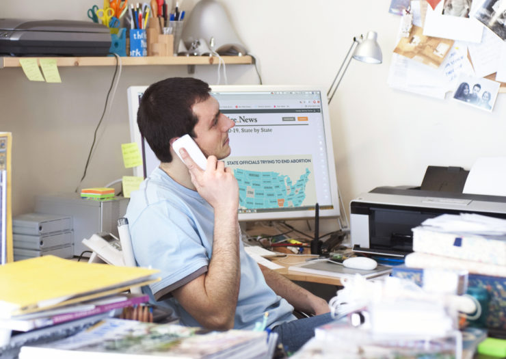 [Photo: A man sitting in front of his computer at a messy desk talks on the phone.]