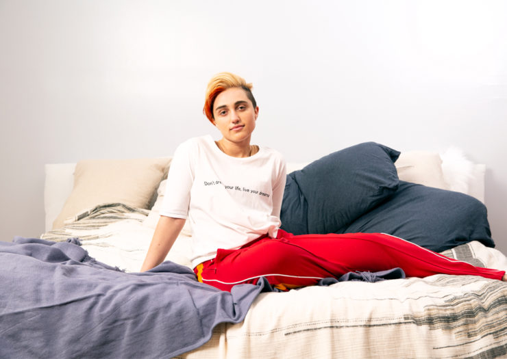[Photo: A gender nonconforming person sits in bed and looks at the camera.]