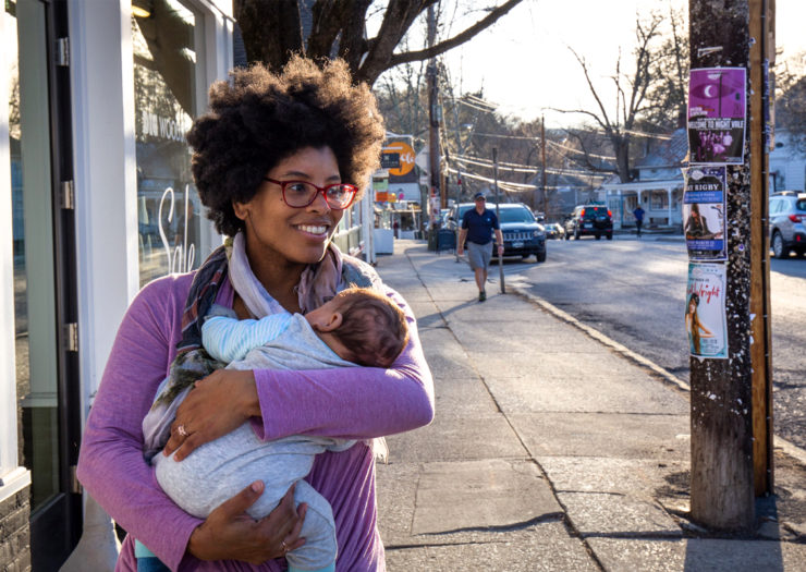 [Photo: A young, Black mother smiles as she walks down the street and holds her baby in her arms.]