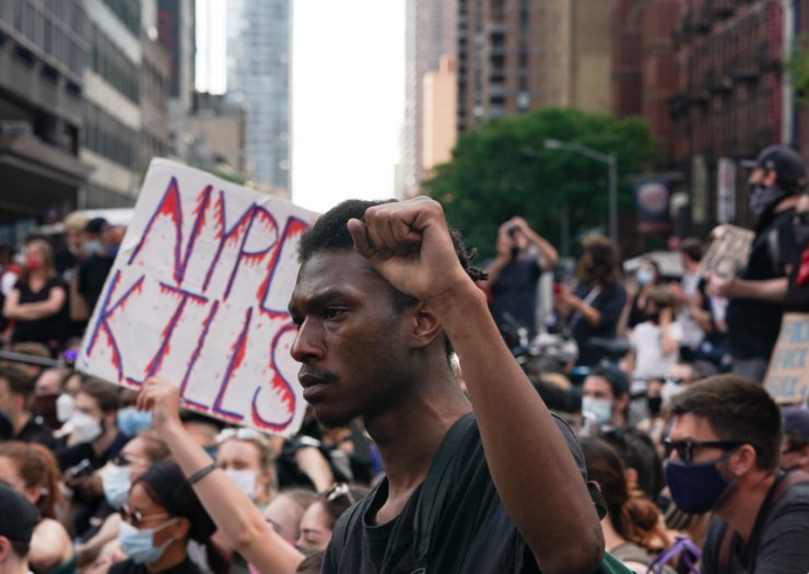 [Photo: A Black man holds his fist up at a police brutality protest, a sign in the background says 'NYPD KILLS'.']