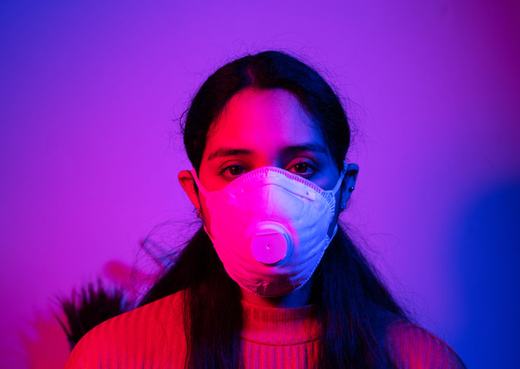 [Photo: A young woman of color wears a mask and looks at the camera sadly. She is illuminated by lights of purple and red hues.]