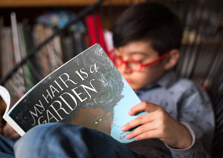 [Photo: A young boy of color wears red glasses and reads a book titled 'My Hair Is A Garden' as he sits in a chair.]