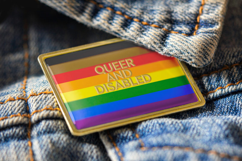 [Photo: A close-up of a denim jacket with a Pride flag pin that reads 'Queer and Disabled.']