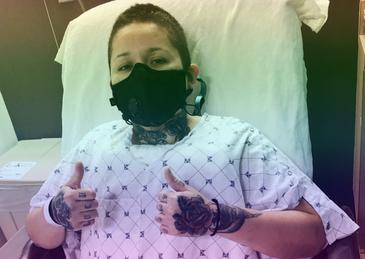 [Photo: A nonbinary person sits in a hospital room post-operation, giving two thumbs up in a hospital gown and protective face mask.]