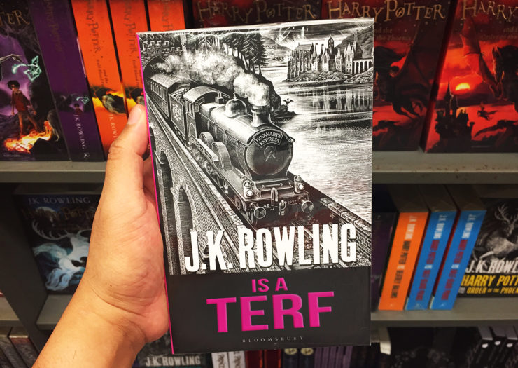 [Photo: A person holds a book by J.K. Rowling up against a bookstand with a myriad of other J.K. Rowling books. The book title is edited to say 'J.K. Rowling is a TERF in magenta letters.]