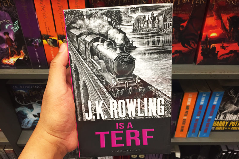 [Photo: A person holds a book by J.K. Rowling up against a bookstand with a myriad of other J.K. Rowling books. The book title is edited to say 'J.K. Rowling is a TERF in magenta letters.]