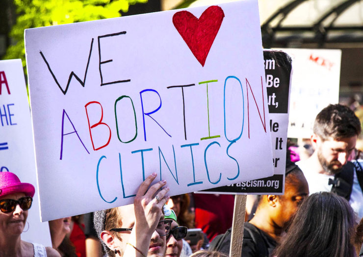 [Photo: A person holds a sign that reads 'We Love Abortion Clinics' during a pro-abortion rally.]
