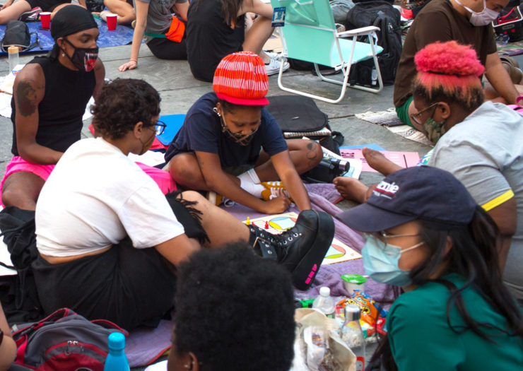 [Photo: A group of protesters of color wearing masks sit in a circle and play a board game.]