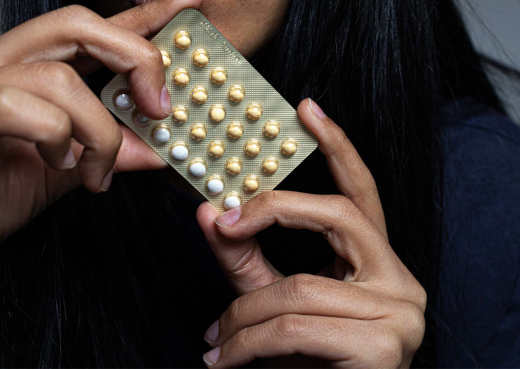 [Photo: A person of color with long, black hair holds a blister pack of birth control pills.]