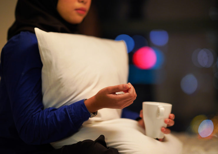 [Photo: A young Muslim person holds a mug in one hand and a pill in the other. She looks hesitant.]
