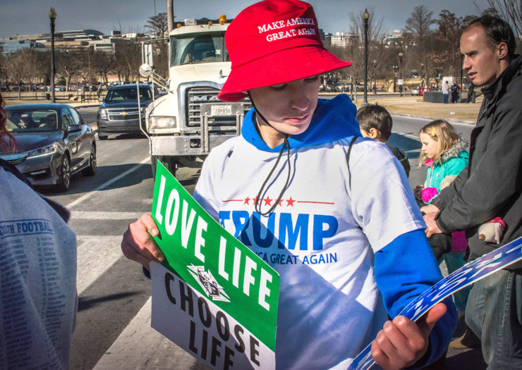 [Photo: A young person wearing a 'Make America Great Again' hat and Trump shirt holds anti-abortion posters as he crosses the street.]
