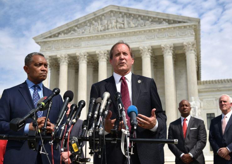[PHOTO: Ken Paxton standing in front of a row of microphones] in front of the Supreme Court building]