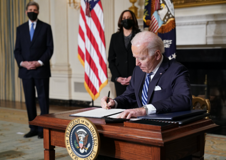 [PHOTO: Joe Biden at desk signing with pen. John Kerry and Kamala Harris stand a distance away]