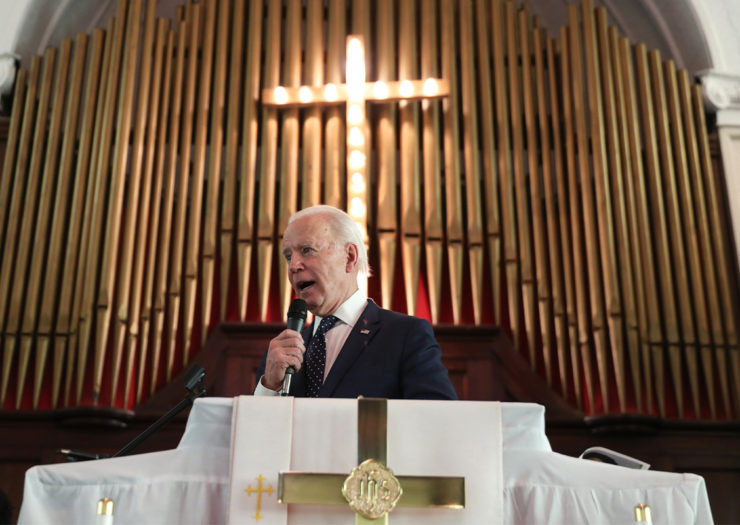[Photo: President Biden speaks at a church. A large, illuminated cross hangs behind him.]