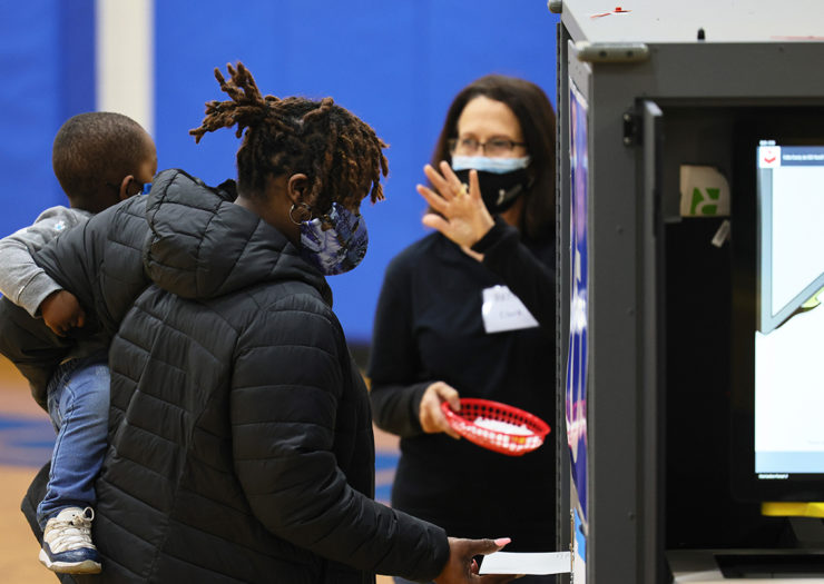 [Photo: A Black mom wearing a mask holds her child as she submits her ballot at a voting booth. Next to her is a white woman wearing a mask and holding up a hand.]