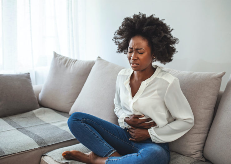[PHOTO: Black woman wearing white top and jeans sitting cross-legged on couch with hands on stomach in pain]