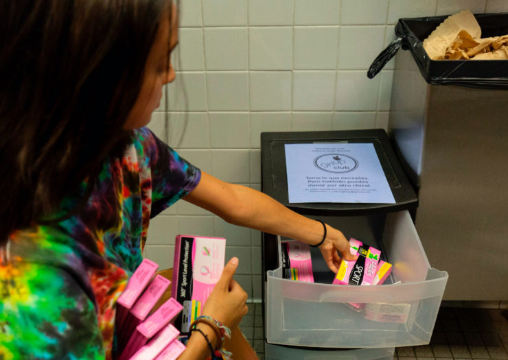 [PHOTO: A teen holds boxes of tampons and stocks a plastic drawer in a bathroom]
