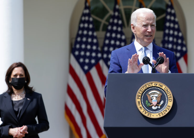 [PHOTO: Kamala Harris wears a dark suit and mask in the background. Joe Biden in a blue suit in the foreground speaking behind a podium]