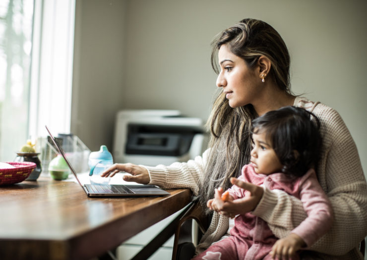 Brown-skinned woman with toddler in her lap as she types on her laptop