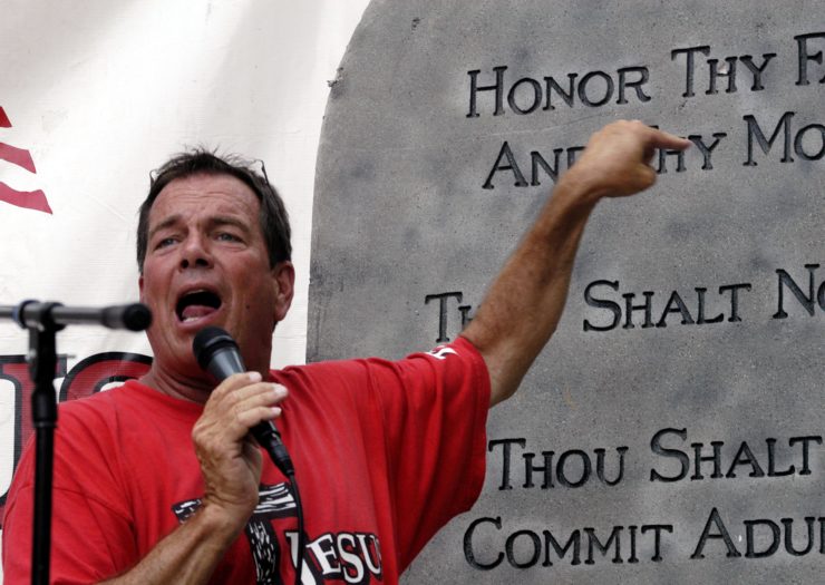 [A man yells into a microphone and points at a large Ten Commandments statue behind him.]