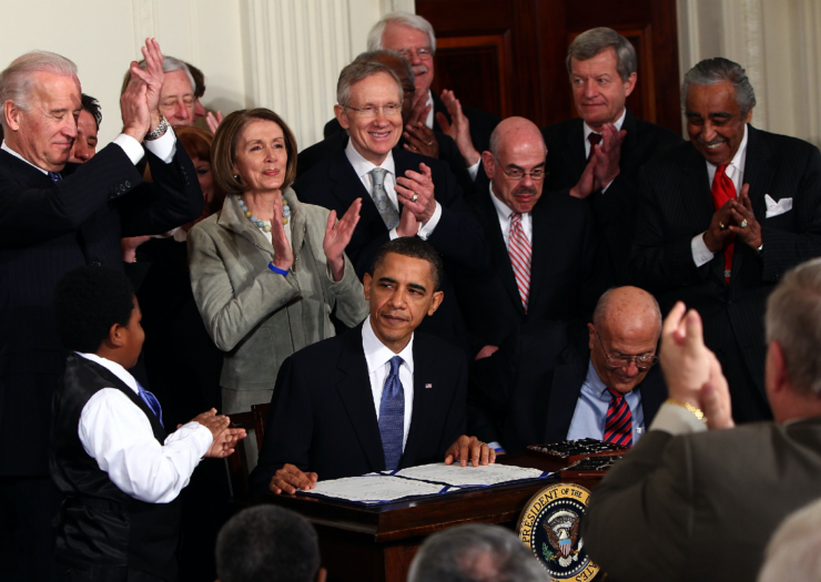 Barack Obama sitting behind a desk with lawmakers surrounding him and cheering