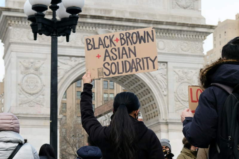 Photo of the back of a woman holding a sign that reads Black brown Asian solidarity